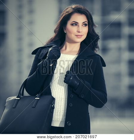 Happy young woman with handbag walking in a city street. Stylish fashion model in dark blue classic coat outdoor