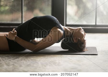 Young attractive woman practicing yoga at home, stretching in Fish exercise, Matsyasana pose, working out, wearing sportswear, black shorts and top, indoor close up image, studio background