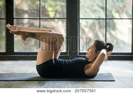 Young woman with ankle tattoo making Abdominal Knee Crunches for upper, lower abs, Sit Ups exercise, working out, wearing sportswear black shorts and top, indoor full length, studio or home background