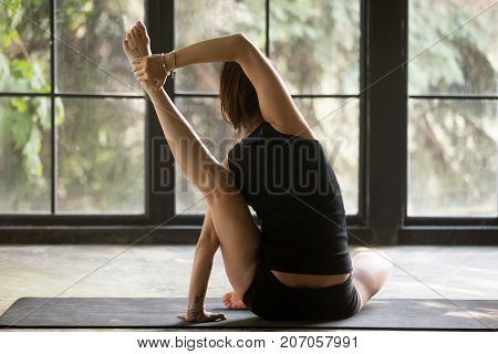 Young attractive woman practicing yoga at home, sitting in Compass exercise, Surya Yantrasana pose, working out, wearing sportswear, black shorts, top, indoor full length, studio background, rear view