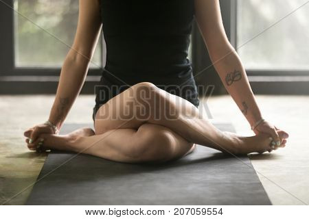 Young sporty woman practicing yoga at home, sitting in Gomukasana exercise, Cow Face pose, working out, wearing sportswear black shorts, top, indoor close up image, studio background. Wellness concept