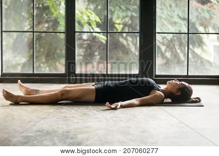 Young attractive woman practicing yoga at home, lying in Savasana exercise, Corpse pose, working out, wearing sportswear, black shorts and top, indoor full length, studio background