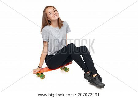 Teen girl in full length sitting on skate board smiling at camera, isolated on white background