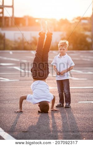 The boy is showing a headstand on the street. Younger brother admires his brother
