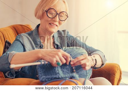 Enjoy the process. Joyful nice aged stylish woman sittiing in the arm chasir and smiling while knitting
