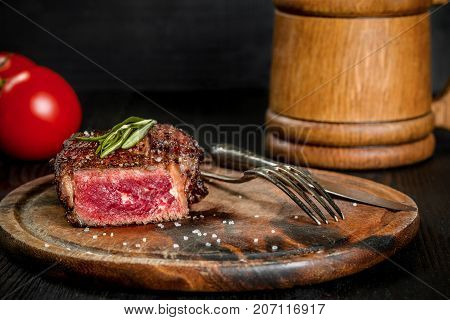 Grilled steak seasoned with spices and fresh herbs served on a wooden board with wooden mug of beer and fresh tomato. Black wooden background. Still life