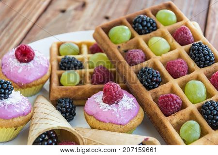Belgian waffels, cupcakes and ice cream cone with grape, blueberry, raspberry on wooden background