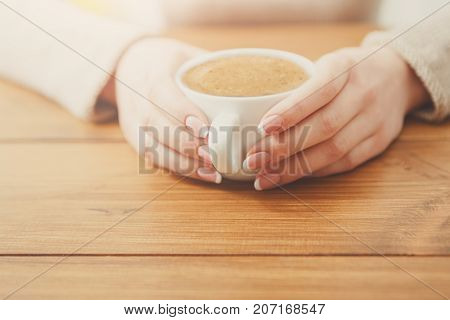 Female holding a cup of coffee while sitting at wooden table, shallow depth of field.
