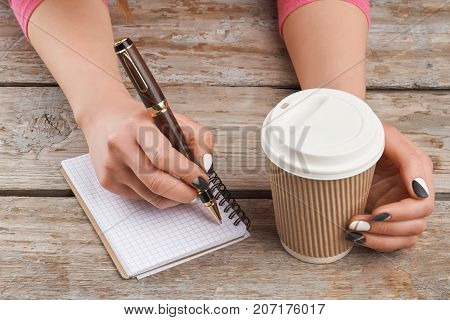 Manicured hand writing a note. Young woman hands holding pen and disposable cup of cappuccino on old wooden background. Female manicured hands, paper notebook, cardboard cup.