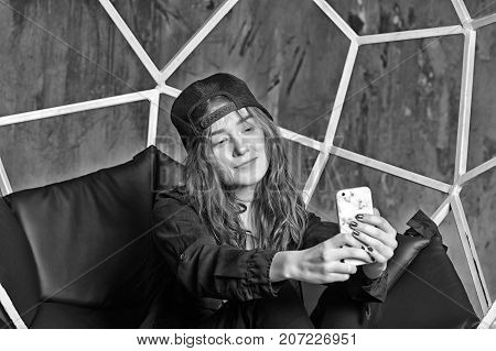 Self expression concept. Girl or woman in baseball cap making selfie with smartphone on brown chair. Technology and mobile device black and white