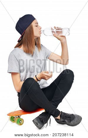 Teen girl in full length sitting on skate board drinking water from a bottle isolated on white background