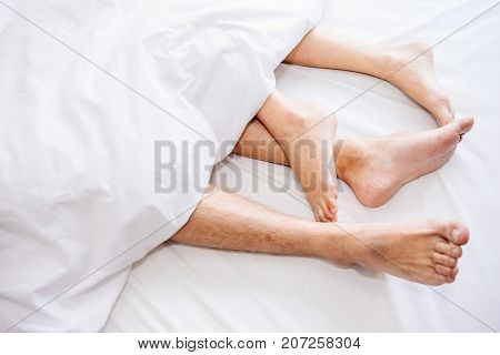 The bare foot of couples sleeping on the bed and cover by the white blanket on a bright Sunday morning.