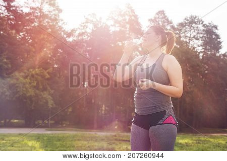 Sweaty overweight woman drinking water after a long workout. Selective focus.