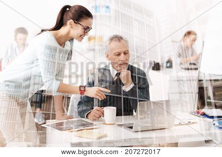Generation bound. Cheerful atrractive young woman talking with her senior colleagues who is sitting at the table and using laptop