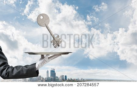 Closeup of waiter's hand in white glove presenting stone key symbol on metal tray with cityscape view on background. 3D rendering.