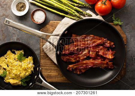 Cooked sizzling hot tasty crispy bacon on a skillet, scrambled eggs and vegetables ready for breakfast overhead shot