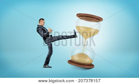 A tiny businessman kicks and shakes a large hourglass with half of its sand already in the lower chamber. Harness your time. Efficient workhours. Fight against time.