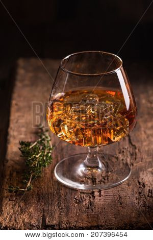 Glass of brandy or cognac with ice on old oak wooden table. Dark photo.