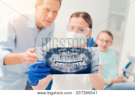 Have a look. Selective focus on a computed tomography scan picture of teen jaws held by a female stomatologist standing next to a male parent and discussing some dental problems of his child.