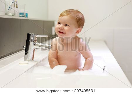 Cute adorable baby taking bath in washing sink and grab water tap. Little healthy girl with big blue eyes having fun and playing with soap foam