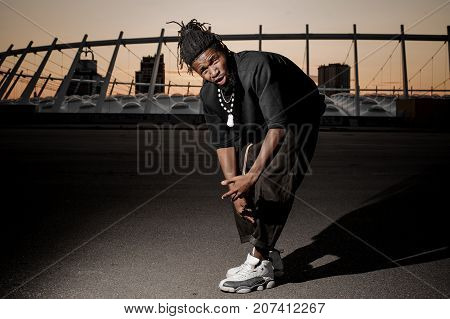 Outdoor full length portrait of the young afro american guy in the half squat dressed in black clothes