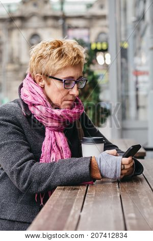 Adult blonde woman sitting with coffee and smartphone in outside cafe on street.