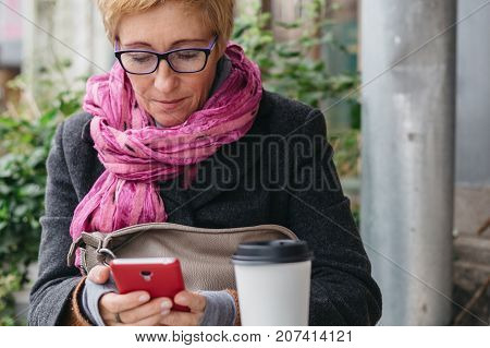 Adult blonde woman browsing smartphone and having drink from paper cup in outside cafe.