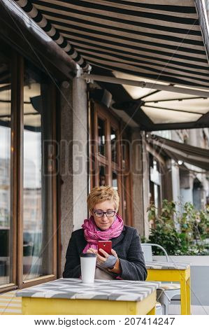Adult woman sitting at table in outside cafe and using smartphone while having coffee.