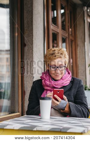 Adult woman sitting at table in outside cafe and using smartphone while having coffee.