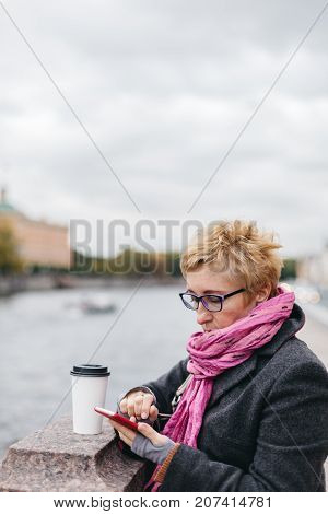 Blonde adult woman standing with coffee cup and using smartphone on waterfront.