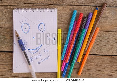 Open white notepad with drawing and inscription BACK TO SCHOOL with colorful felt-tip pens and ball pens on the wooden table