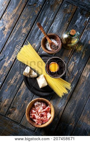 Ingredients for traditional italian pasta alla carbonara. Uncooked spaghetti pancetta bacon parmesan cheese egg yolk salt pepper in olive wood bowls over old plank background. Top view with space