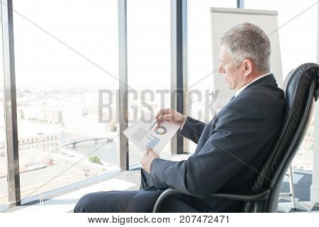 Mature businessman sitting alone in office with large panoramic windows with view at city and looking at diagrams and financial reports