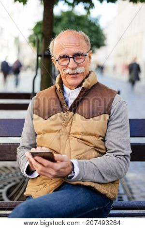 Enjoying A Beautiful Day. Cheerful Mature Man Sitting On Wooden Bench With Phone Typing And Smiling.