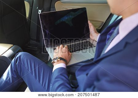 Close-up of a young businessman who uses a laptop to work in the backseat of a car.