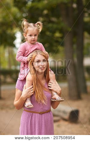 Close- up of a small daughter sitting by her mother on her shoulders strollimg in the park.