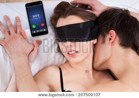Close-up Of A Man In Bed With Blindfolded Woman And Wife Calling On Smart Phone