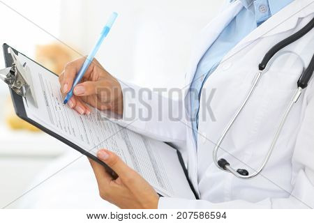 Close-up of a female doctor filling  up medical form at clipboard while standing straight in hospital.