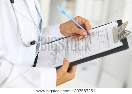 Close-up of a female doctor filling  up medical form at clipboard while standing straight in hospital.