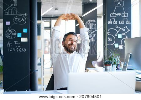 Handsome young businessman in his office, sitting at the desk, stretching his arms.