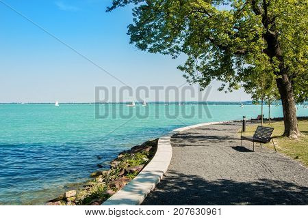 A view of Balaton lake with white yachts at the horizon and trees bench and footpath at the foreground Tihany Hungary.