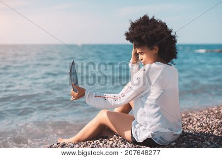 Wide-angle view of young black woman sitting on a wicker chair outdoors in street modern cafe nearshore and swiping on the screen of digital tablet while waiting for her order sunny summer day