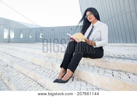 Thoughtful young woman enjoying literature and sitting on stairs in campus. Concentrated Hispanic businesswoman learning management. Business school concept