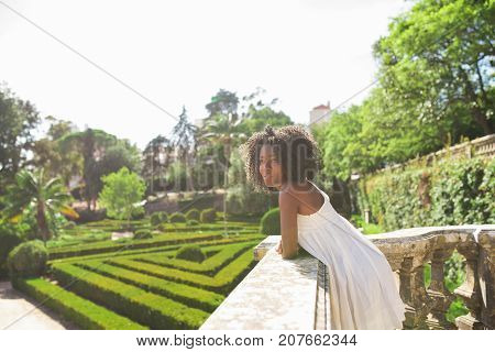 Closeup portrait of dreamy young attractive African American woman standing and leaning on stone railing in park with beautiful garden in background. Side view.