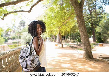 Portrait of happy young African-American woman wearing white blouse and carrying backpack walking in park, looking at camera and smiling. City break and travel concept
