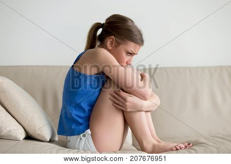Sad depressed young woman sitting on sofa embracing her knees, upset heartbroken teen girl feeling vulnerable ashamed, frustrated adolescent teenager having problem, suffering from bullying or abuse