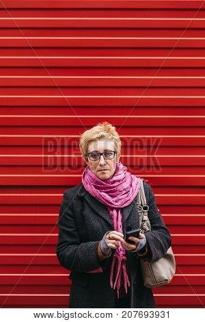 Adult woman using smartphone standing at red fence on city street.