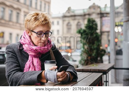 Adult blonde woman sitting in outside cafe with paper cup and surfing smartphone.