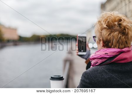 Back view of adult blonde woman taking smartphone shots from esplanade.
