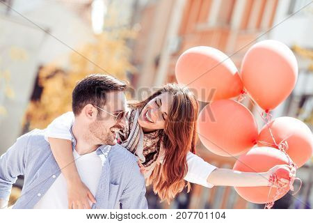Romantic couple having fun while walking in the city. Girl is sitting piggyback and holding balloons.